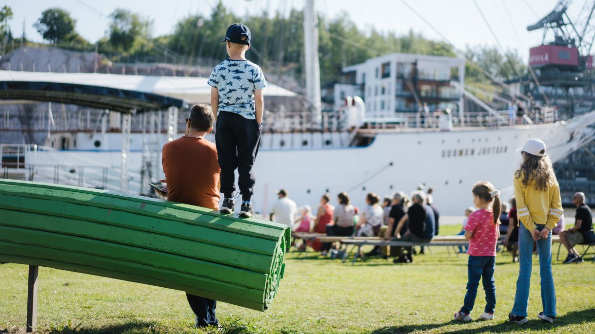 Ihmisiä istumassa penkeillä katsomassa isoa purjelaivaa. Lapsi seisoo penkillä