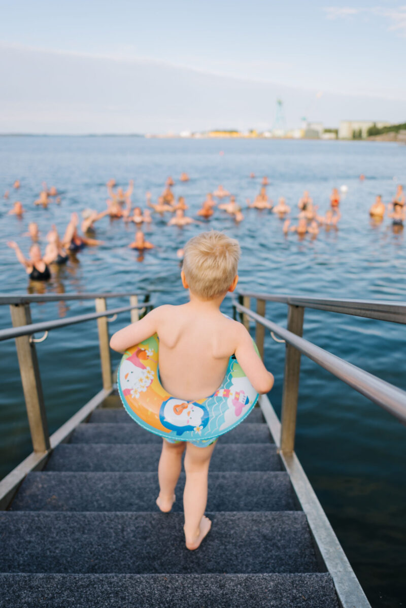 Pieni lapsi selin kuvattuna, värikkään uimarenkaan kanssa laskeutumassa laiturin portaista kohti avomerta, jossa suuri joukko ihmisiä uiskentelee ja heilauttaa käsiään taustalla näkyvän sataman ja nostureiden horisontin edessä.