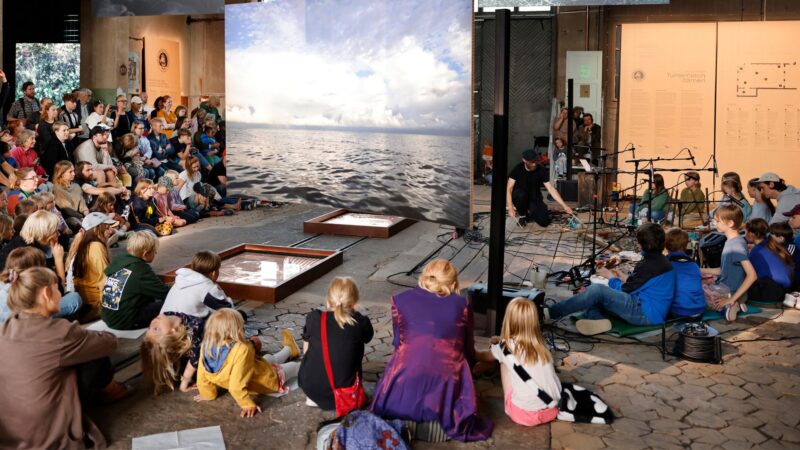 Audience of children and adults seated on the floor and benches attentively watching a live performance in an indoor space with a large backdrop image of the sea.
